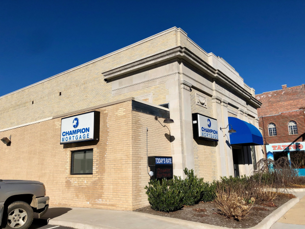 Ein zweistöckiges Gebäude mit vernagelten Fenstern, einem geparkten Auto, Pflanzen und Bäumen unter einem bewölkten Himmel, mit "Champion Mortgage, Inc."-Schildern.