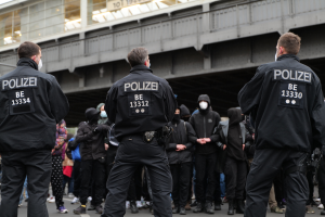 Eine Gruppe von Polizeibeamten in Uniform steht vor einer Menge von Menschen in schwarzen Uniformen und Masken, mit einer Brücke und einem Gebäude im Hintergrund, während einer Demonstration in einer Stadt.