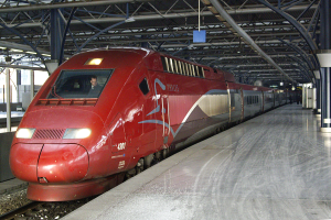 Ein roter Hochgeschwindigkeitszug steht auf einem Bahnhof mit einer Plattform auf der rechten Seite, einige Menschen stehen und im Hintergrund sind Säulen, Stangen und andere Gegenstände zu sehen.