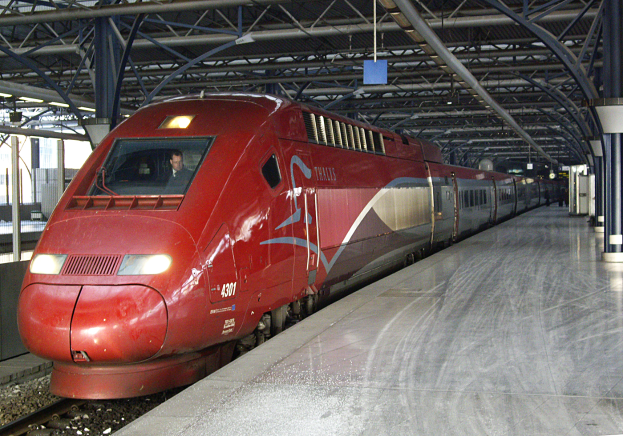 Ein roter Hochgeschwindigkeitszug steht auf einem Bahnhof mit einer Plattform auf der rechten Seite, einige Menschen stehen und im Hintergrund sind Säulen, Stangen und andere Gegenstände zu sehen.