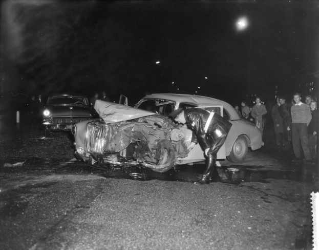 Schwarz-weiß-Foto eines auf der Straße stehenden, verungl√∫ckten Autos mit mehreren Personen in der Nähe und beleuchtetem Hintergrund.