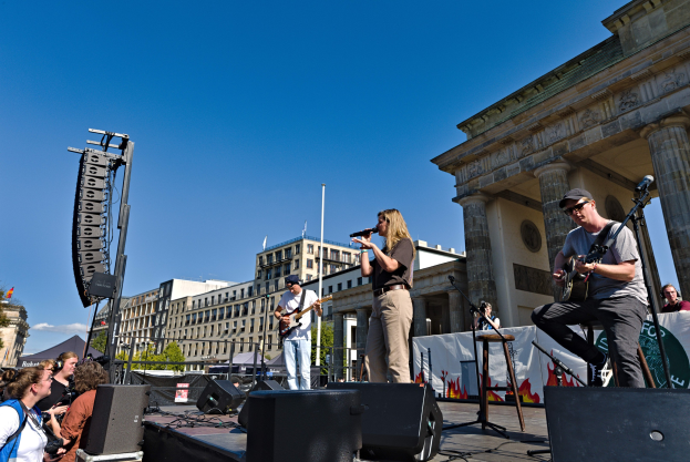 Gruppe von Menschen auf der Bühne mit Instrumenten und Mikrofonen vor dem Brandenburger Tor, mit Lautsprechern und Equipment im Bild, vor Gebäuden, Bäumen und einem klaren Himmel.