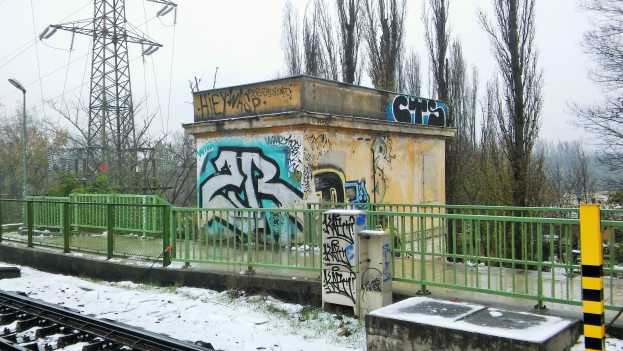 Eisenbahnschiene mit Graffiti, umgeben von Zäunen, Pfölen, Lichtern und Bäumen, führend zu Törmen mit Dröhnen unter einem klaren Himmel, mit Schnee am Boden.