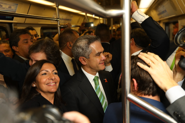 Ein Mann im Anzug und Krawatte steht neben einer Frau in einem überfüllten U-Bahn-Wagen, umgeben von mehreren Menschen mit Kameras und einem Innenraum voller Stangen und Lichter.