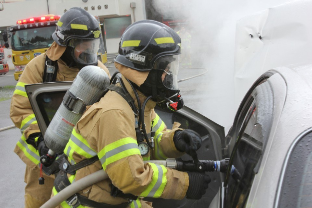 Zwei Feuerwehrleute in Schutzausrütstüchern verwenden einen Schlauch, um ein brennendes Auto zu löschen, mit Rauch und Fahrzeugen und einem Gebäude im Hintergrund.