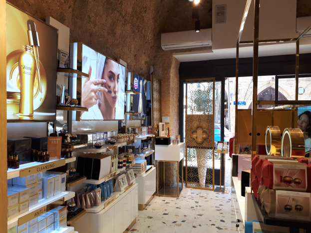 Innenansicht eines Kosmetikladens mit verschiedenen Produkten auf Regalen, an der Wand montierten Bildschirmen, Deckenleuchten, einer Klimaanlage und Glastüren, die eine Person, ein Auto und ein Gebäude draußen zeigen.