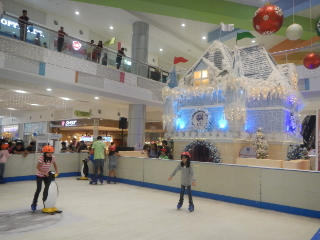 Menschen, die auf einer Eisbahn in einem Einkaufszentrum Schlittschuhlaufen, umgeben von festlichen Dekorationen, mit anderen, die auf Geländern und Säulen stehen.