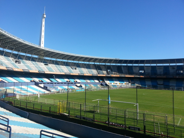 Großes Stadion mit einem Fußballfeld, das von einem Zaun umgeben ist, einem Turm im Hintergrund und einem klaren blauen Himmel.