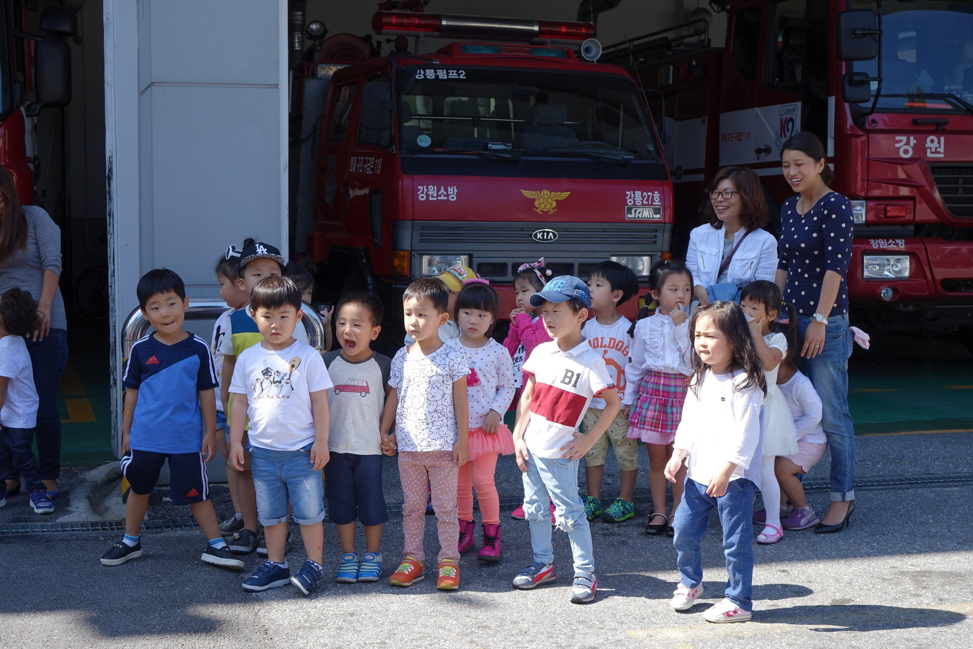 Eine Gruppe von Kindern vor mehreren Feuerwehrautos auf einem Feuerwehrahof, einige tragen Mützen.