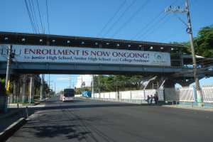Ein Bus fährt unter einer Brücke mit einem Schild, auf dem "Jetzt Anmeldung" steht, während Menschen auf dem Gehweg darunter gehen, mit Strommasten und Drähten an der Straße und Bäumen und einem klaren blauen Himmel im Hintergrund.