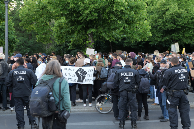 Eine große Gruppe von Menschen nimmt an einer Black Lives Matter Demonstration auf einer Straße in Berlin teil, einige halten Schilder und andere tragen Mützen und Taschen, während im Vordergrund ein Fahrrad und im Hintergrund Bäume und ein Pfahl zu sehen sind.