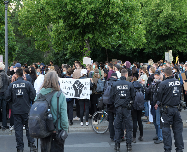 Eine große Gruppe von Menschen nimmt an einer Black Lives Matter Demonstration auf einer Straße in Berlin teil, einige halten Schilder und andere tragen Mützen und Taschen, während im Vordergrund ein Fahrrad und im Hintergrund Bäume und ein Pfahl zu sehen sind.