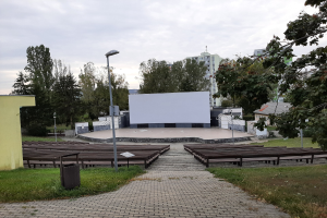 Outdoor movie theater in a park with benches, grass, a trash bin, light poles, trees, buildings, and a cloudy sky.