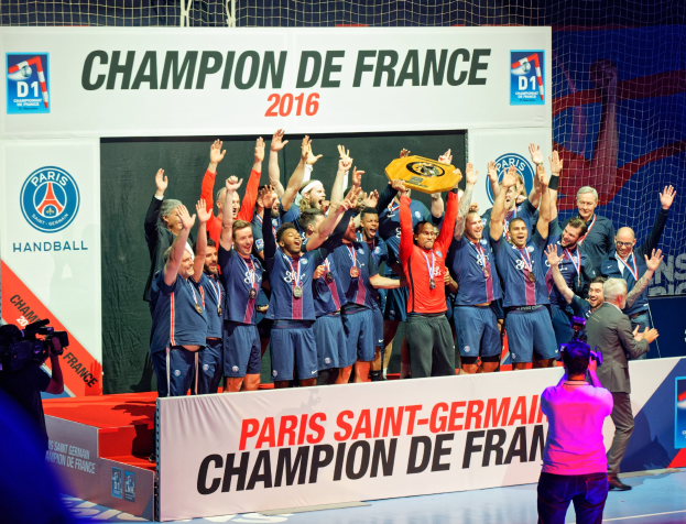Eine Gruppe von Männern auf einer Bühne, die eine Trophäe halten, während jemand sie filmt; im Hintergrund ist ein "Champion de France 2016"-Schild zu sehen.