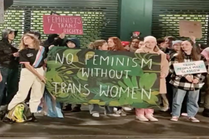 Gruppe von Menschen mit einer Transparent haltend, auf dem "Kein Feminismus ohne Trans-Frauen" steht und Schilder in öffentlichem Raum mit einer Wand und Rollladen im Hintergrund.