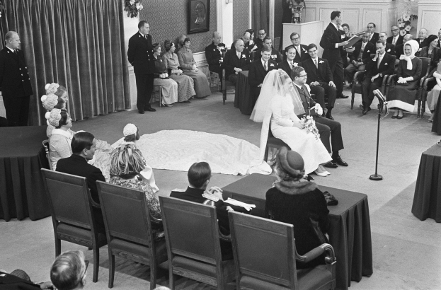 Schwarzes und weißes Foto einer Hochzeitszeremonie mit sitzenden und stehenden Menschen, einem Mann mit Mikrofon, einer Frau mit Blumenstrauß, einem Podium mit Mikrofon auf der rechten Seite, Blumengestecken und einem Vorhang auf der linken Seite, einem Rahmenfoto und einer Tür im Hintergrund.