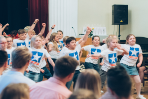 Eine Gruppe von Kindern in weißen Hemden und blauen Shorts tanzt auf einer Bühne vor einem sitzenden Publikum, mit einem Vorhang, Lautsprecher und einem Tisch mit einem Mikrofon im Hintergrund.