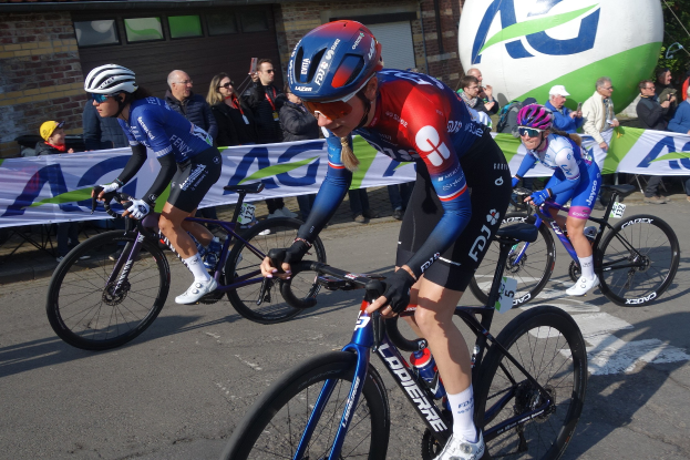 Gruppe von Radfahrern fährt eine Straße entlang, mit einem Banner im Hintergrund, einige tragen Helme und halten Kameras, mit einem Gebäude, Bäumen und einem Ballon im Hintergrund.