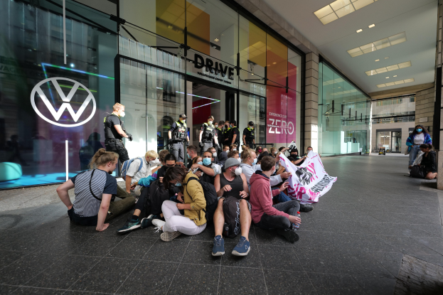 Eine Gruppe von Menschen sitzt vor einem Volkswagen-Autohaus auf dem Boden, einige halten ein Transparent, mit sichtbaren Glaswänden und Logo des Autohauses.