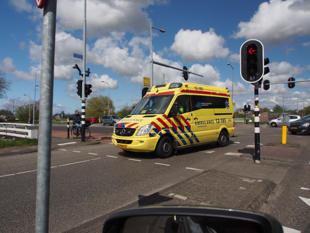 Ein Rettungswagen fährt auf einer Straße neben einer Ampel, mit einem Radfahrer im Vordergrund und städtischen Elementen wie Gebäuden, Bäumen und Laternen im Hintergrund.