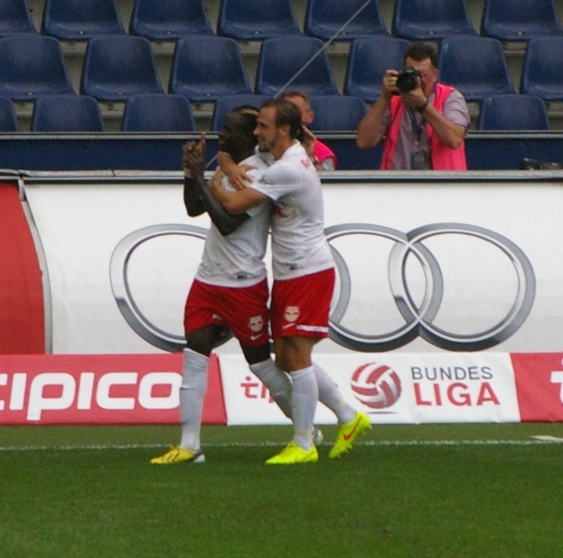Zwei Fußballspieler feiern ein Tor auf einem Feld mit einer Menge, einem Fotografen, einem Banner, Stühlen und einer Wand im Hintergrund.
