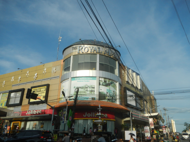 Eine belebte Stadtstraße mit fahrenden Fahrzeugen, Fußgängern auf dem Gehweg und einem großen Gebäude mit der Aufschrift "Royal Mall" im Hintergrund, umgeben von parkenden Autos, Bäumen, Strommasten und Drähten unter einem Himmel.