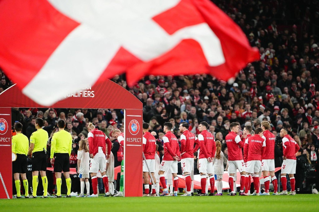 Gruppe von Menschen auf einem Fußballfeld mit einer roten und weißen Flagge im Vordergrund, einem Bogen mit dem Text "Bayern Munich vs. Bayern Munich Wetten & Vorschau" im Hintergrund und einer großen Menge im Stadion.