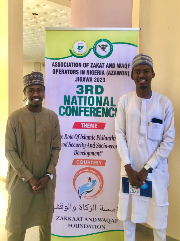 Zwei Männer stehen nebeneinander vor einem Banner mit der Aufschrift "3. Nationales Konferenz" mit einer Säule und Deckenbeleuchtung im Hintergrund.