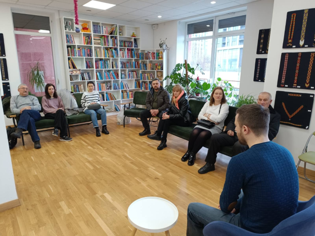 Gruppe von Menschen, die um einen Tisch in einem Raum mit Regalen, Pflanzen und Deckenleuchten sitzen und sich bei einem Gesundheit und Wohlfühl-Workshop in Berlin unterhalten.