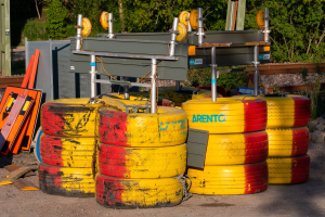 Ein Haufen gelber und roter Reifen auf einem Schmutzfeld umgeben von verstreuten Metallobjekten, Brettern und anderem Schutt, mit Bäumen, Pflanzen und einer Hütte im Hintergrund.