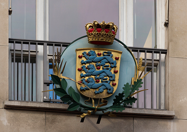 Wappen der britischen Königsfamilie an einer Gebäudewand mit einer Krone und -Blatt-Logo, Fenstern und Metallgittern im Hintergrund.