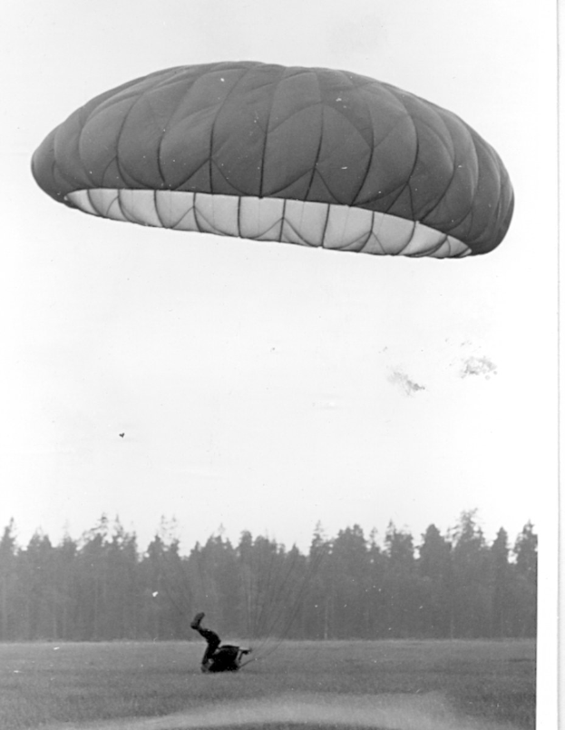 Schwarz-weißes Foto einer Person beim Paragleiten in der Luft mit einem Fallschirm, umgeben von Bäumen und einem klaren Himmel, mit Text unten.