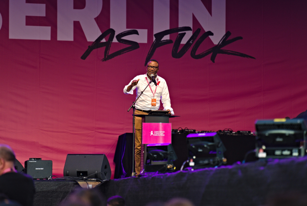 Mann auf einer Bühne vor einem Publikum mit einem Mikrofon, weißes Hemd und braune Hose tragend, mit einem pinken Banner 'Berlin as Fuck' im Hintergrund.