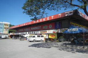 Ein öffentlicher Markt mit einem vor ihm geparkten Van, umgeben von Gebäuden, Geschäften, Schildern, Fahrzeugen, Menschen, die unter einem Zelt sitzen, Geländern, Bäumen und einem klaren blauen Himmel.