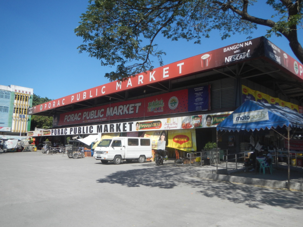 Ein öffentlicher Markt mit einem vor ihm geparkten Van, umgeben von Gebäuden, Geschäften, Schildern, Fahrzeugen, Menschen, die unter einem Zelt sitzen, Geländern, Bäumen und einem klaren blauen Himmel.