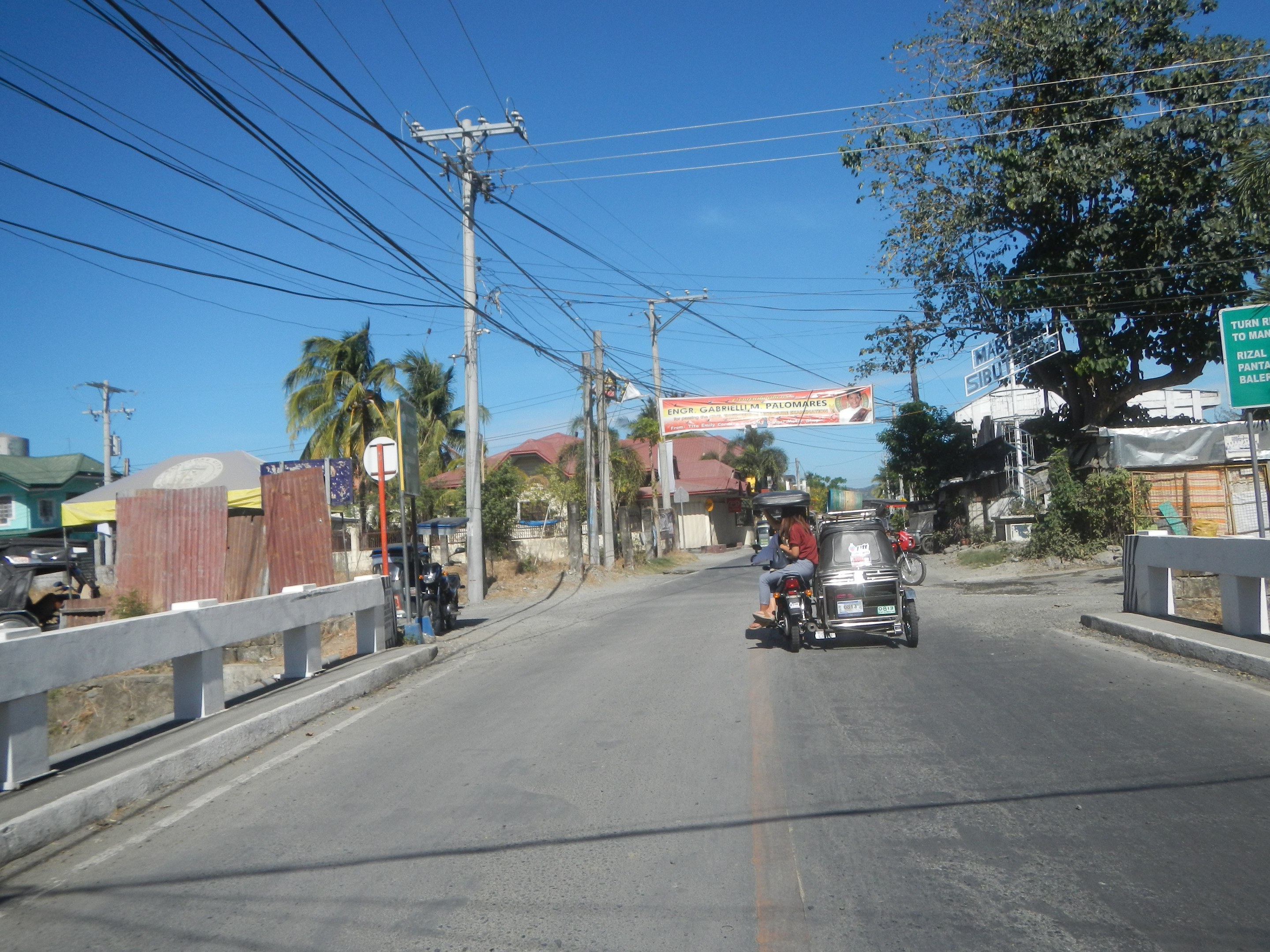 Eine Person, die ein Motorrad auf einer Straße mit Strommasten, Drähten, Geländern, Bäumen, Gebäuden und einem klaren blauen Himmel im Hintergrund fährt.