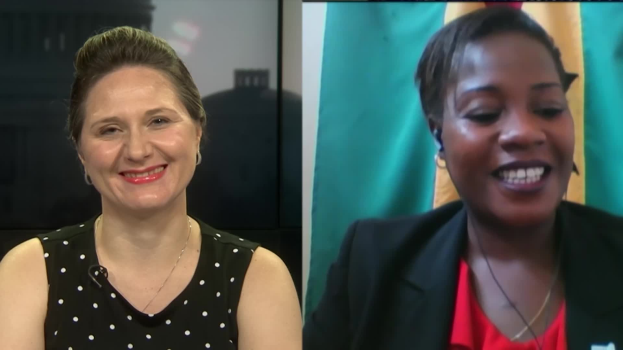 Zwei Frauen, die lächeln, waßren bei einem Videoanruf, eine in einem schwarzen und weißen Kleid, die andere in einer unbestimmten Farbe, mit einer Fahne an der Wand hinter ihnen in einer öffentlichen Einrichtung.