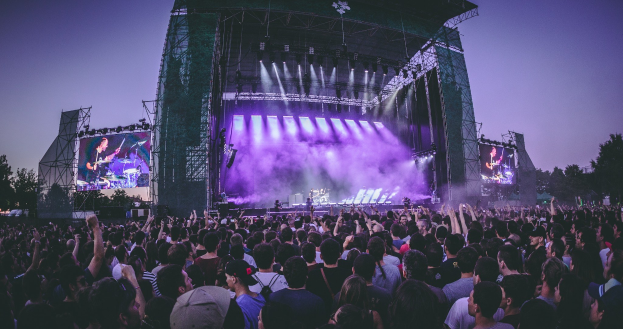 Eine große Menschenmenge auf einem Musikfestival schaut auf einer hell erleuchteten Bühne mit Instrumenten und Bildschirmen, die vor einem Gebäude, Bäumen und einem klaren blauen Himmel aufgebaut sind.