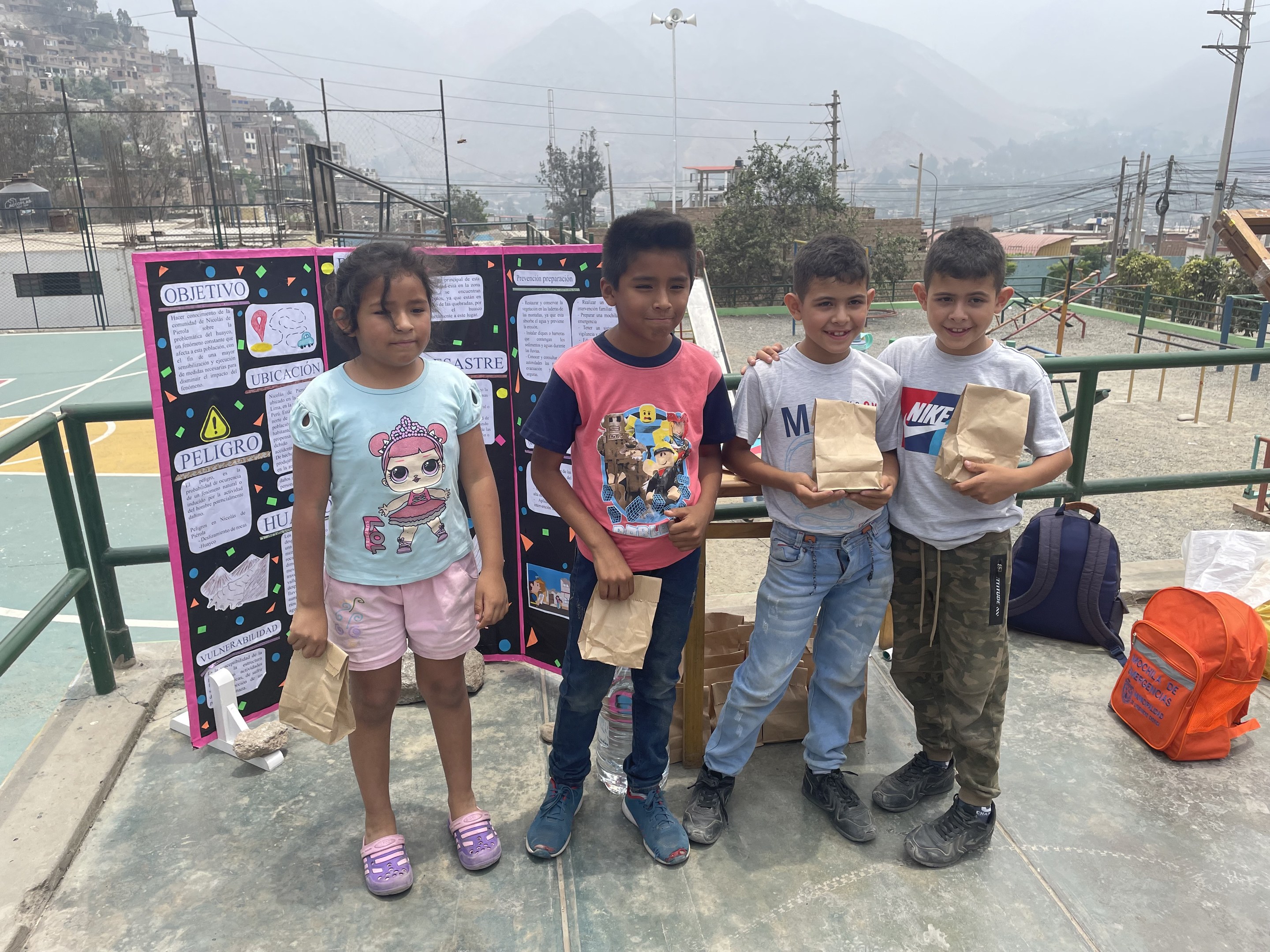 Gruppe von Kindern auf einem Basketballfeld mit Papiertüten in den Händen, mit Tüten und Gegenständen rechts und einer Tafel mit Text links, vor einem Hintergrund aus Zaun, Pfosten, Straßenlaternen, Bäumen, Gebäuden, Hügeln und Himmel.
