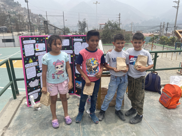 Gruppe von Kindern auf einem Basketballfeld mit Papiertüten in den Händen, mit Tüten und Gegenständen rechts und einer Tafel mit Text links, vor einem Hintergrund aus Zaun, Pfosten, Straßenlaternen, Bäumen, Gebäuden, Hügeln und Himmel.