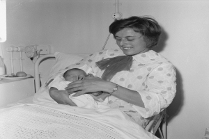 Eine Frau in einem Krankenhausbett hält ein Neugeborenes im Arm, umgeben von einem Kissen und einer Decke, mit einem Tisch links daneben und Rohren im Hintergrund.