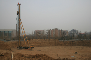 Eine Baustelle mit einem zentralen Kran, umgeben von Gebäuden, Bäumen und einem klaren blauen Himmel, mit Erde bedecktem Boden und verstreuten Gegenständen.