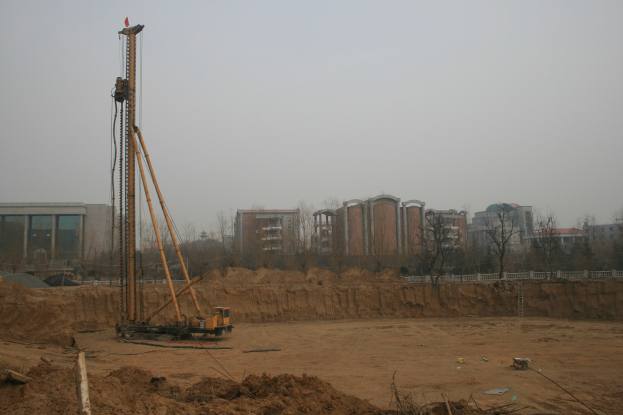 Eine Baustelle mit einem zentralen Kran, umgeben von Gebäuden, Bäumen und einem klaren blauen Himmel, mit Erde bedecktem Boden und verstreuten Gegenständen.