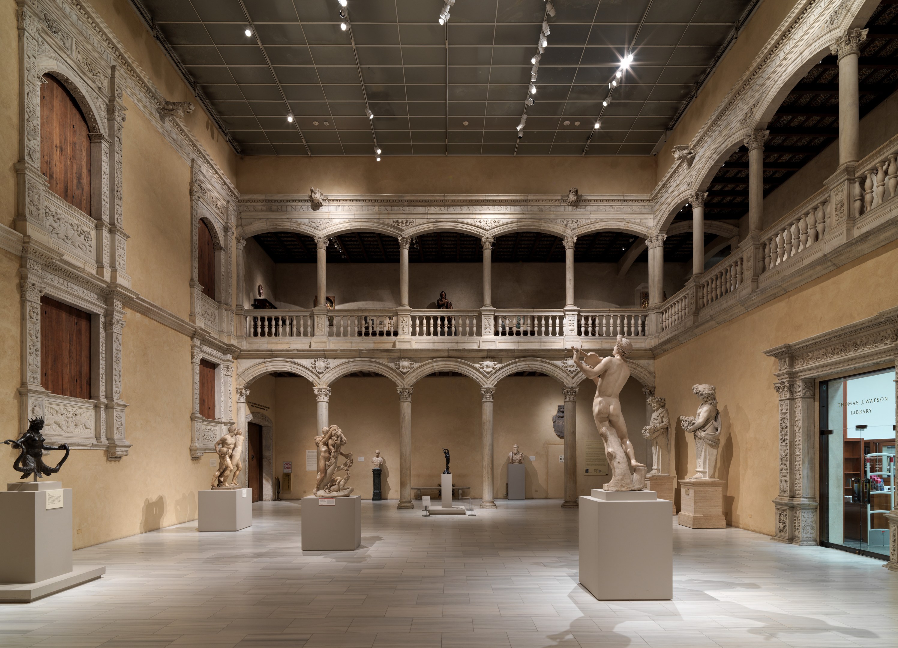 Eine große Museumsgalerie mit zahlreichen Statuen und Skulpturen auf Sockeln, architektonischen Elementen wie Säulen, Geländern und einer beleuchteten Decke sowie einigen Besuchern, die die Kunstwerke betrachten.