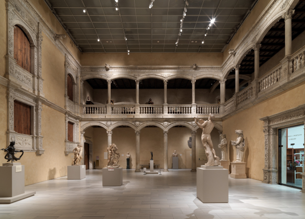 Eine große Museumsgalerie mit zahlreichen Statuen und Skulpturen auf Sockeln, architektonischen Elementen wie Säulen, Geländern und einer beleuchteten Decke sowie einigen Besuchern, die die Kunstwerke betrachten.