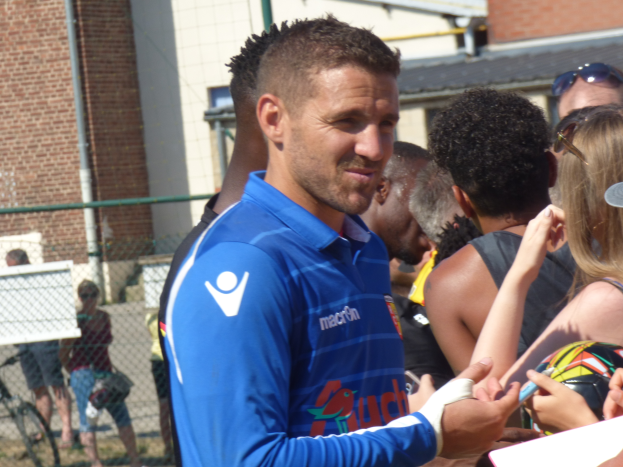 Ein Mann in einem blauen Hemd signiert Autogramme für eine Gruppe von Menschen, einige tragen Mützen und Schutzbrillen, mit einem Gebäude, Maschendrahtzaun und einem Fahrrad im Hintergrund.