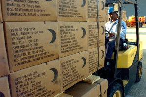 Ein Mann in einem blauen Hemd und einer Mütze fährt einen mit Kartons beladenen Stapler auf einer Straße mit anderen Fahrzeugen und einem klaren blauen Himmel im Hintergrund.