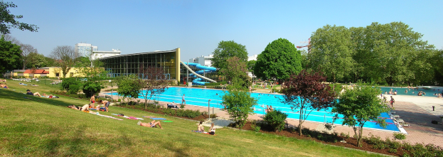 Großer Freibadpool mit Menschen darin und drumherum, umgeben von grünem Gras, Bäumen, Gebäuden und einem klaren blauen Himmel.