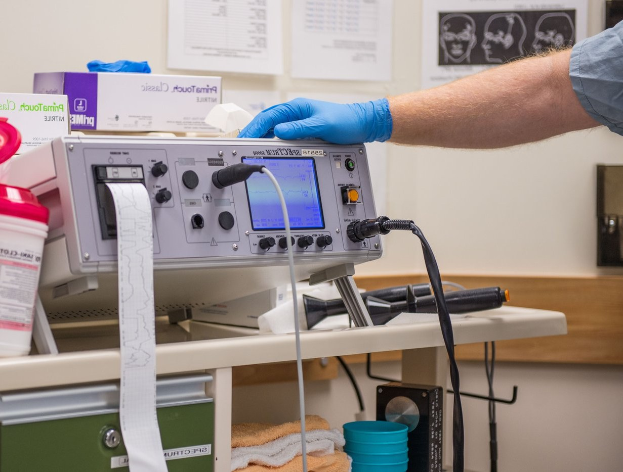 Eine Person in blauen Handschuhen arbeitet an einem medizinischen Gerät auf einem Tisch in einem unordentlichen Krankenhauszimmer mit Kisten, Tassen und Papieren an der Wand.