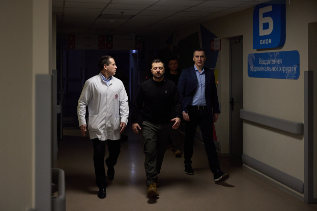 Eine Gruppe von Männern in Not auf einem Krankenhausflur mit Deckenbeleuchtung, einer Tür an der rechten Wand und Texttafeln an der linken Seite.